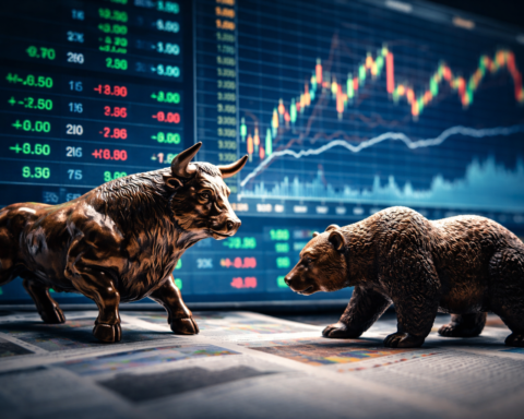 Bronze bull and bear figures facing each other in front of a glowing stock market display with multicolored price charts.