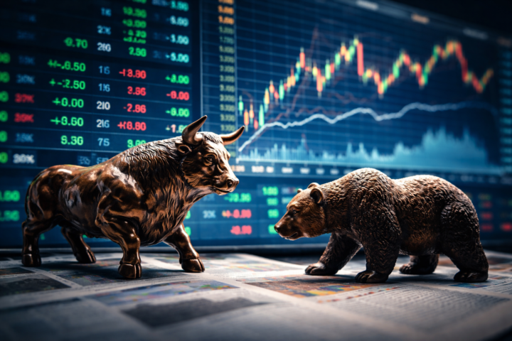 Bronze bull and bear figures facing each other in front of a glowing stock market display with multicolored price charts.