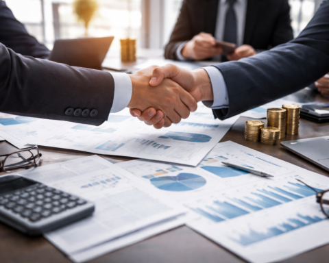 Two business executives shake hands across a conference table covered with financial charts, a laptop, and stacked coins in a modern office.