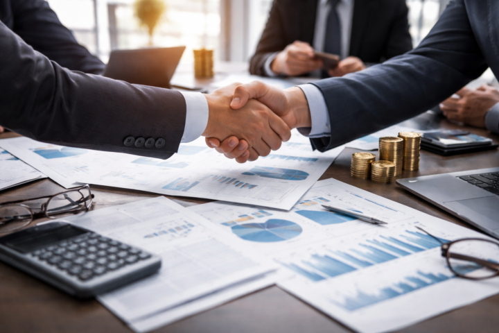 Two business executives shake hands across a conference table covered with financial charts, a laptop, and stacked coins in a modern office.
