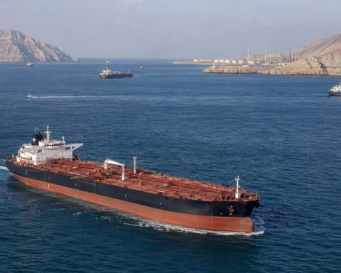 Large crude oil tanker sailing through the Strait of Hormuz with other vessels, rugged Gulf coastline and oil infrastructure in the background.