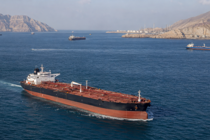 Large crude oil tanker sailing through the Strait of Hormuz with other vessels, rugged Gulf coastline and oil infrastructure in the background.
