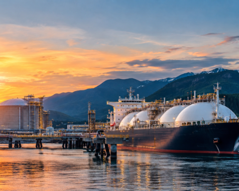 A large LNG tanker docked at a modern liquefied natural gas export terminal on Canada's west coast, with storage facilities, industrial infrastructure, and mountain landscapes in the background at sunset.