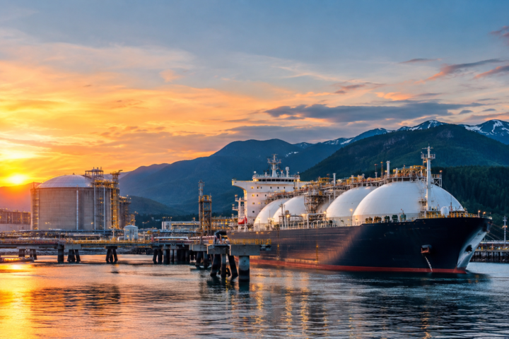 A large LNG tanker docked at a modern liquefied natural gas export terminal on Canada's west coast, with storage facilities, industrial infrastructure, and mountain landscapes in the background at sunset.